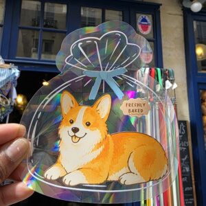 Sticker attrape-soleil holographique en forme de sachet de pain, avec un corgi roux et blanc, tenu à la main devant une boutique.