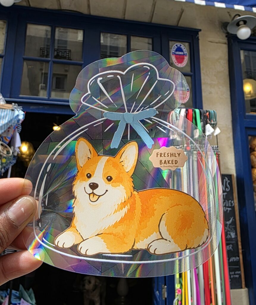 Sticker attrape-soleil holographique en forme de sachet de pain, avec un corgi roux et blanc, tenu à la main devant une boutique.
