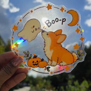 Sticker attrape-soleil holographique représentant un corgi orange et blanc touchant le nez d'un petit fantôme, avec citrouilles, étoiles et lune