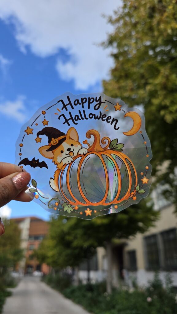 Sticker attrape-soleil holographique Halloween avec un corgi en chapeau de sorcière derrière une citrouille, photographié à la lumière du jour